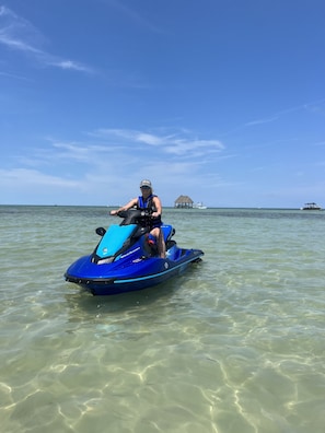 A person on a blue jet ski in clear shallow water with a thatched-roof structure and a boat in the background.