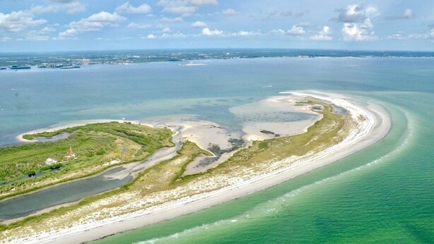 Vue aérienne d’une zone côtière avec une plage de sable, des eaux cristallines et une petite île surmontée d’un phare.