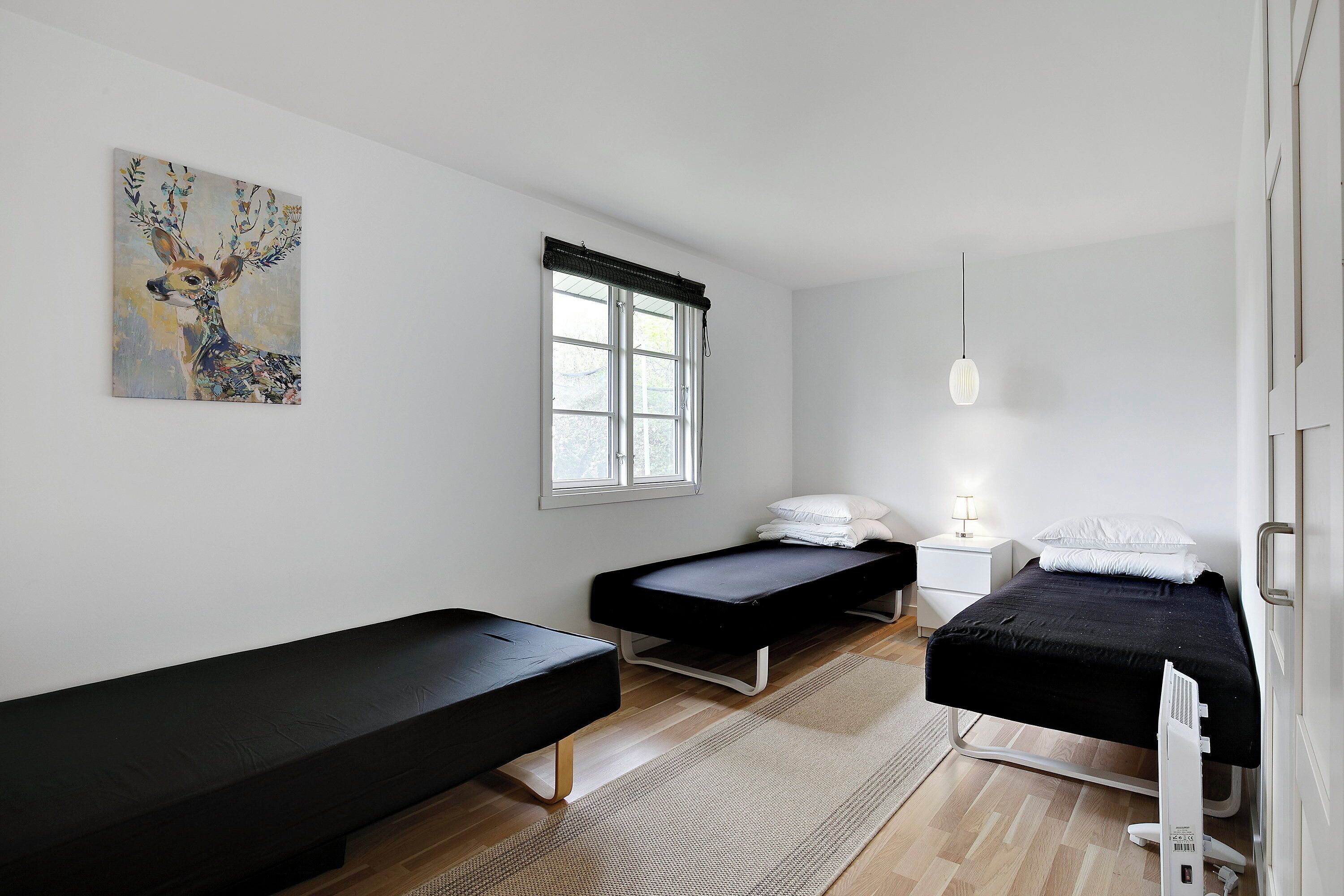 Un dormitorio minimalista con dos camas, una ventana con persianas, una mesita de noche con una lámpara y un cuadro en la pared.