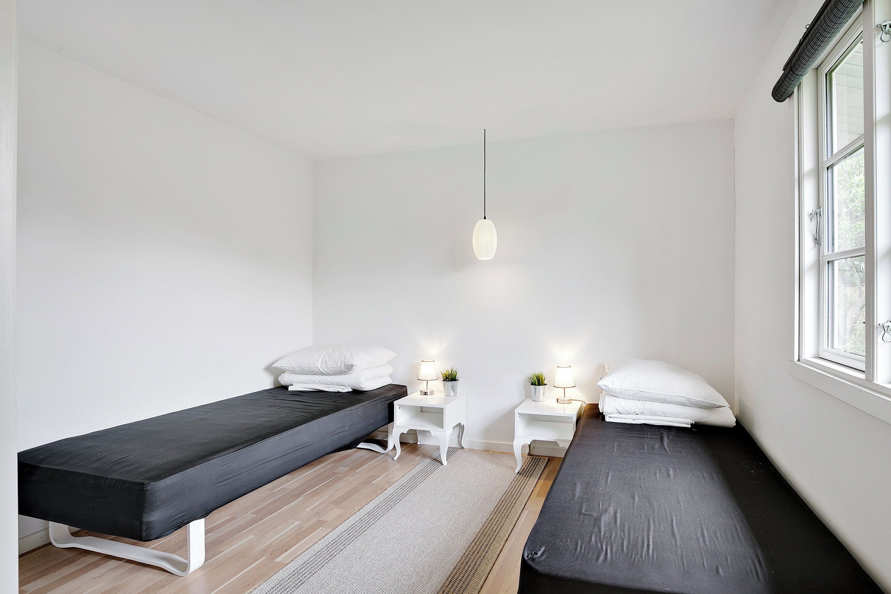 Een kamer met twee bedden, een klein wit nachtkastje met een lamp, een hanglamp en een raam.