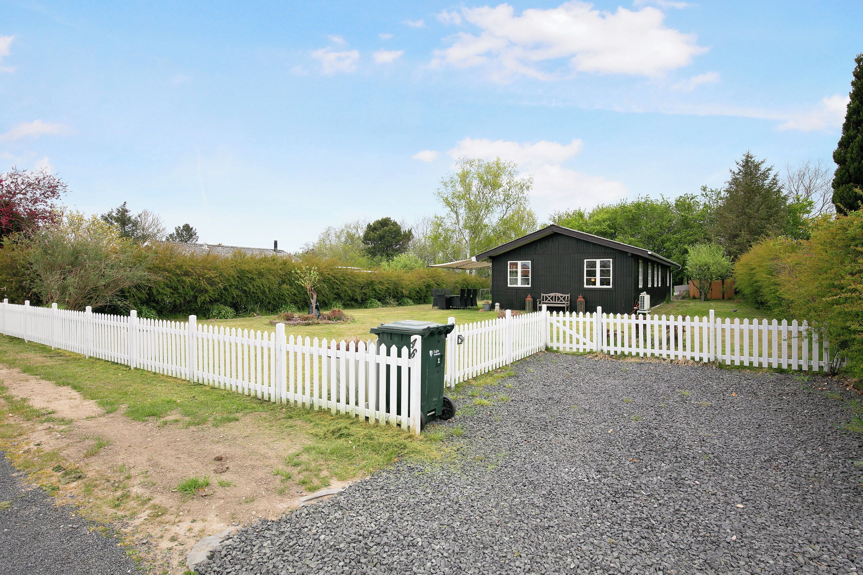 A house with a white picket fence, gravel driveway, and a small green trash bin.