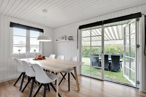 A dining area with a wooden table, white chairs, and a large window with a view of the outdoors. - Lovely cottage for family and friends (Kirke Hyllinge)