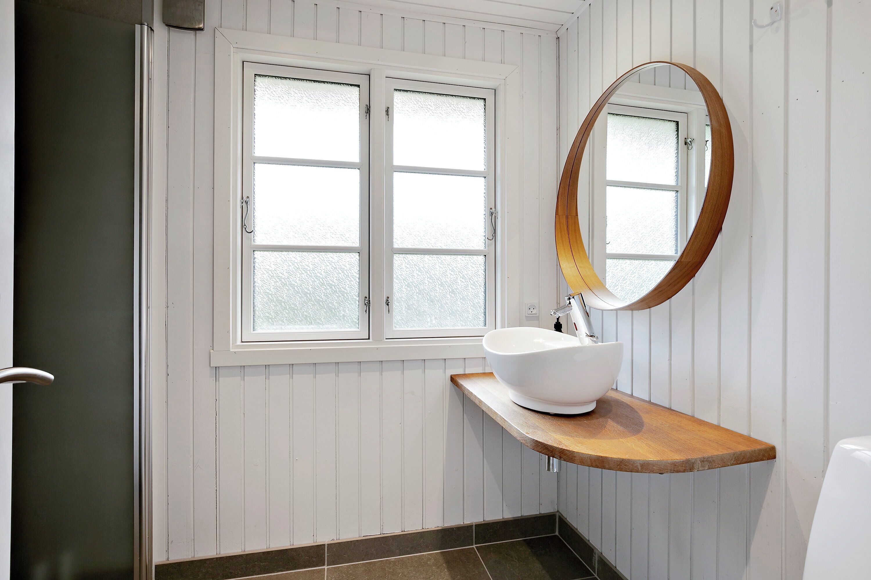 Ein Badezimmer mit einem weißen Waschbecken, einem Holz-Waschtisch, einem runden Spiegel und einem Milchglasfenster.