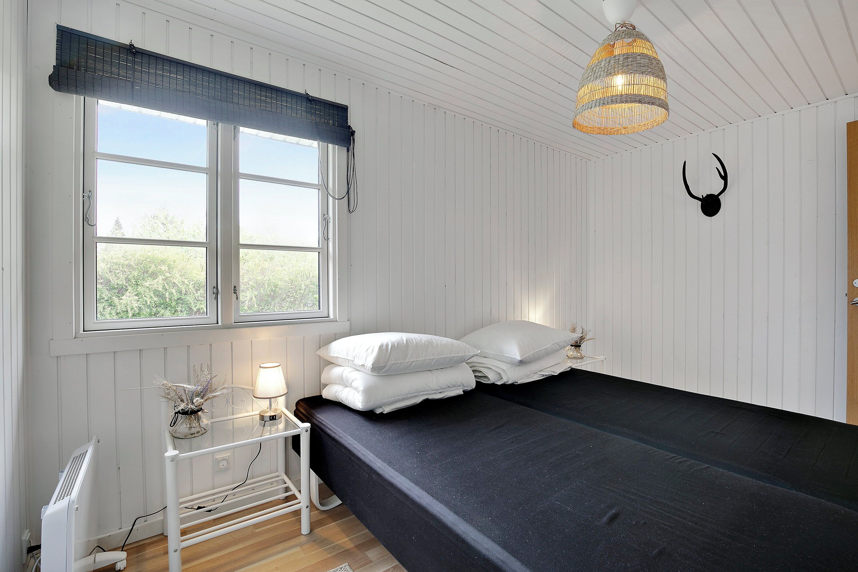 A room with a bed, a desk with a lamp, a window with blinds, and a decorative wall piece.