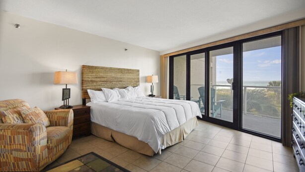 A hotel room with a large bed, a chair, a nightstand, a lamp, and a view of the ocean through a sliding glass door. - Sandestin Beachside II Oasis (Miramar Beach)