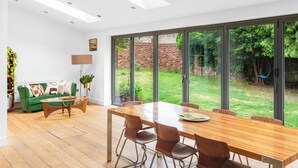 A modern dining area with a wooden table and chairs, a green sofa, and a large window overlooking a garden. - Stunning 3-Bed House in Saffron Walden (Saffron Walden)