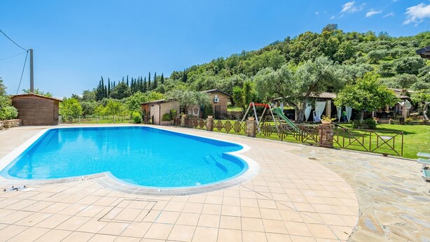 A swimming pool with a tiled deck, surrounded by a fence and houses, set against a hillside. - Local Stone Cottage With Shared Pool, Casal Velino, Italy (Casal Velino)