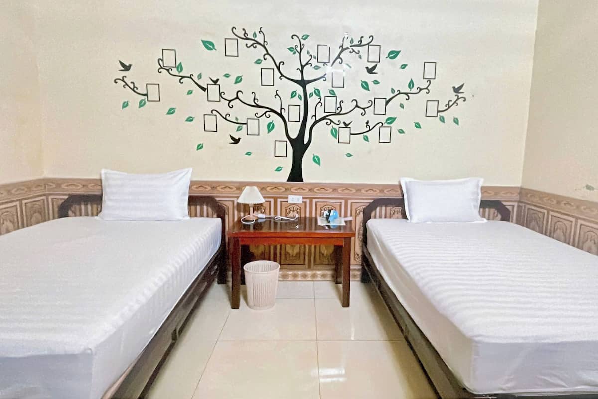 Una habitación de hotel con dos camas individuales, una mesita de noche de madera, una lámpara y un adorno en la pared que representa un árbol con hojas y ramas.