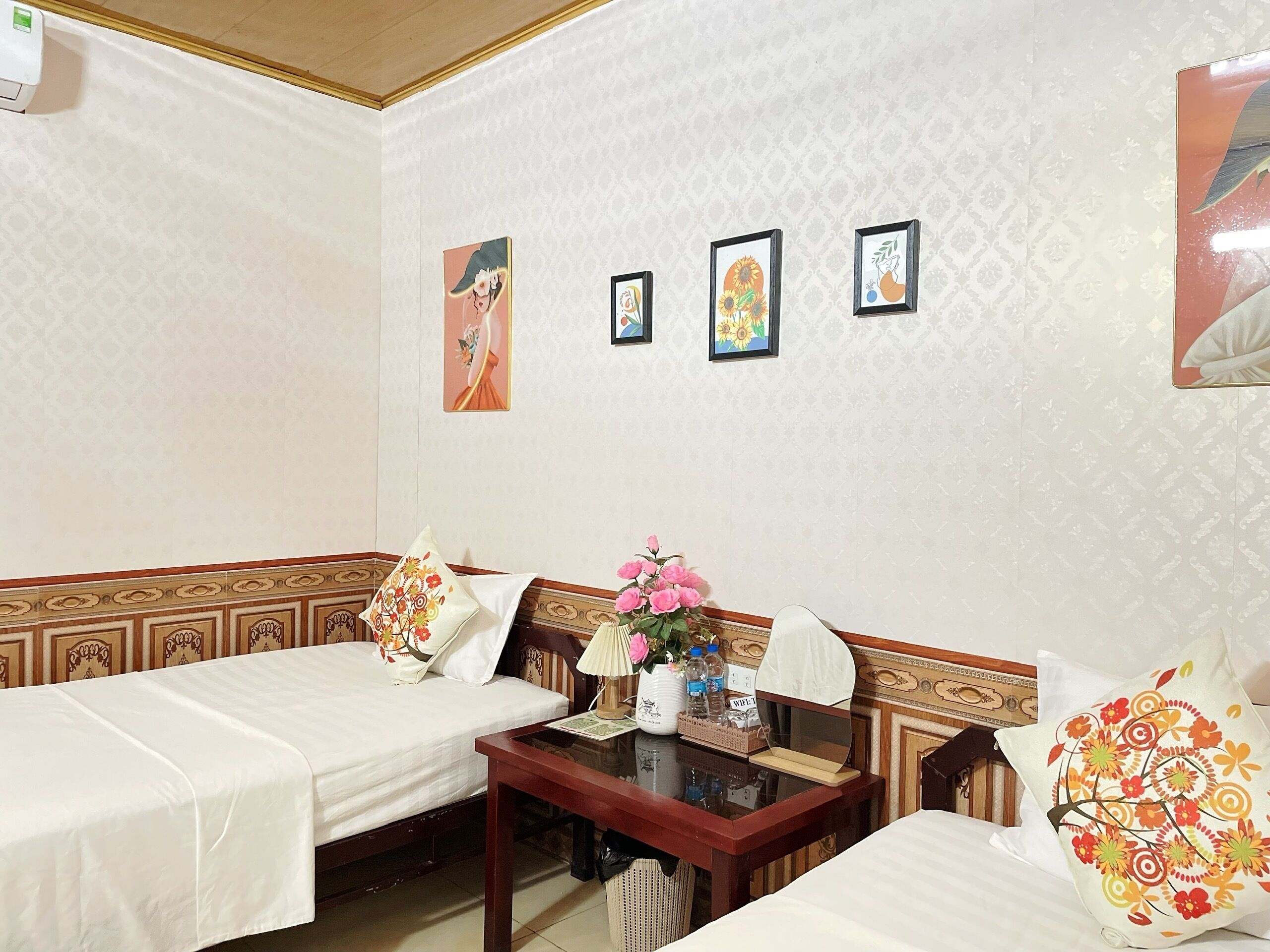 A hotel room with two single beds, a small table with a vase of flowers, and a wall with three framed pictures.