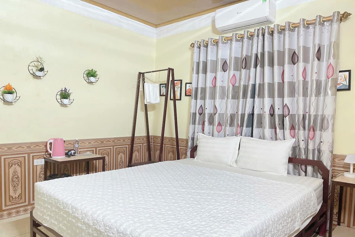 Una cama bien tendida con una colcha blanca, una mesita de noche con una taza rosa y una botella de agua, un aire acondicionado instalado en la pared y una cortina con estampado de hojas.