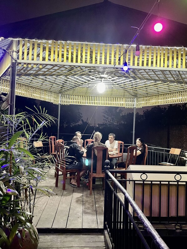Un patio al aire libre con sillas y mesas de madera, una zona de estar cubierta y plantas en macetas.