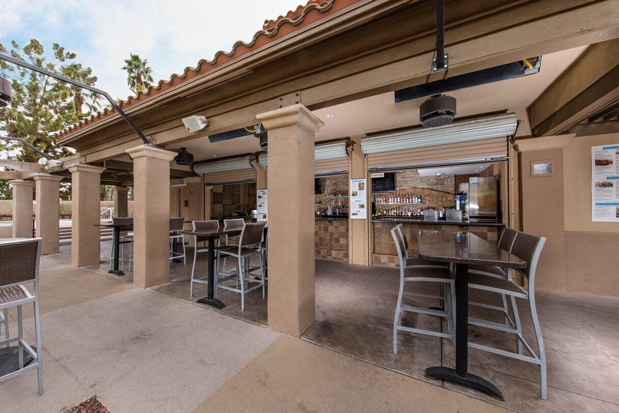 An outdoor bar area with tables and chairs, a counter with beverages, and a stone wall in the background.