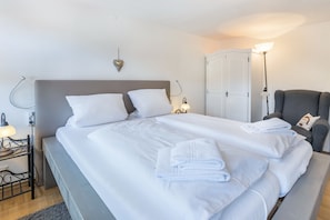 A neatly made bed with white linens and towels, a bedside lamp, and a chair with a cushion.