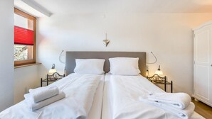 Cama doble con ropa y toallas blancas, lámparas de noche, decoración de pared con forma de corazón y una ventana con persianas.