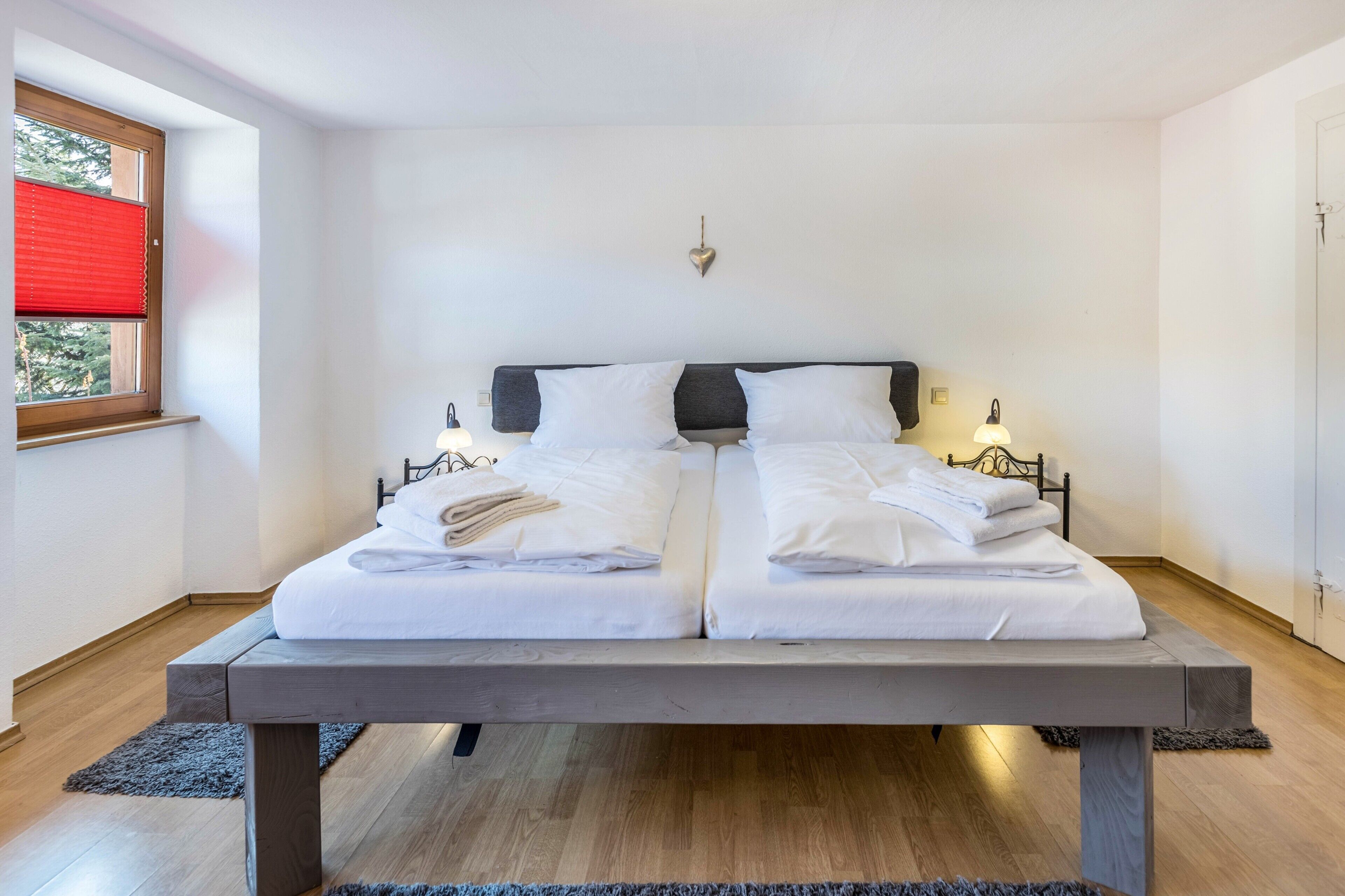 Cama doble con sábanas blancas, cabecera gris, lámparas de noche y una ventana con persiana roja.