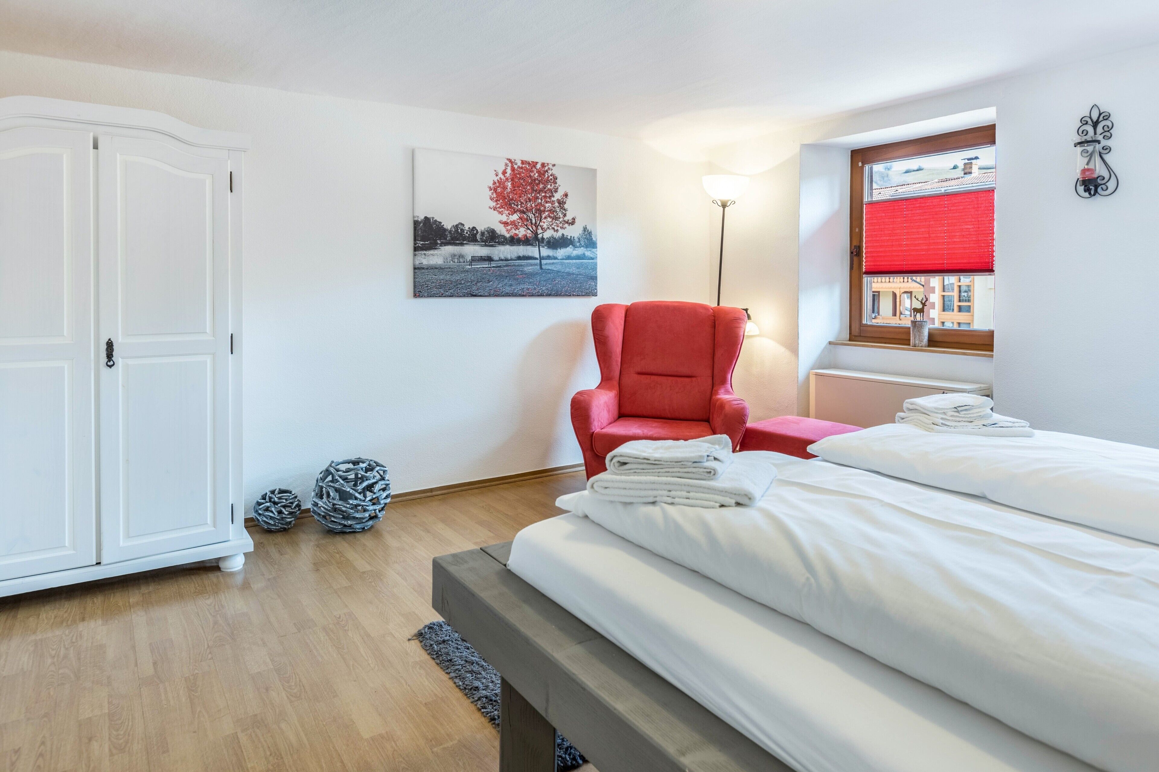 A bedroom with a bed, a red chair, a white wardrobe, a lamp, and a window with a red blind.