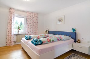 A bedroom with a bed, a nightstand, a window with curtains, a radiator, and a wall-mounted picture.