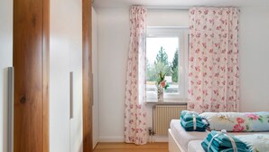 Een slaapkamer met een houten deur, een raam met bloemen gordijnen, een radiator en netjes opgevouwen handdoeken op het bed.