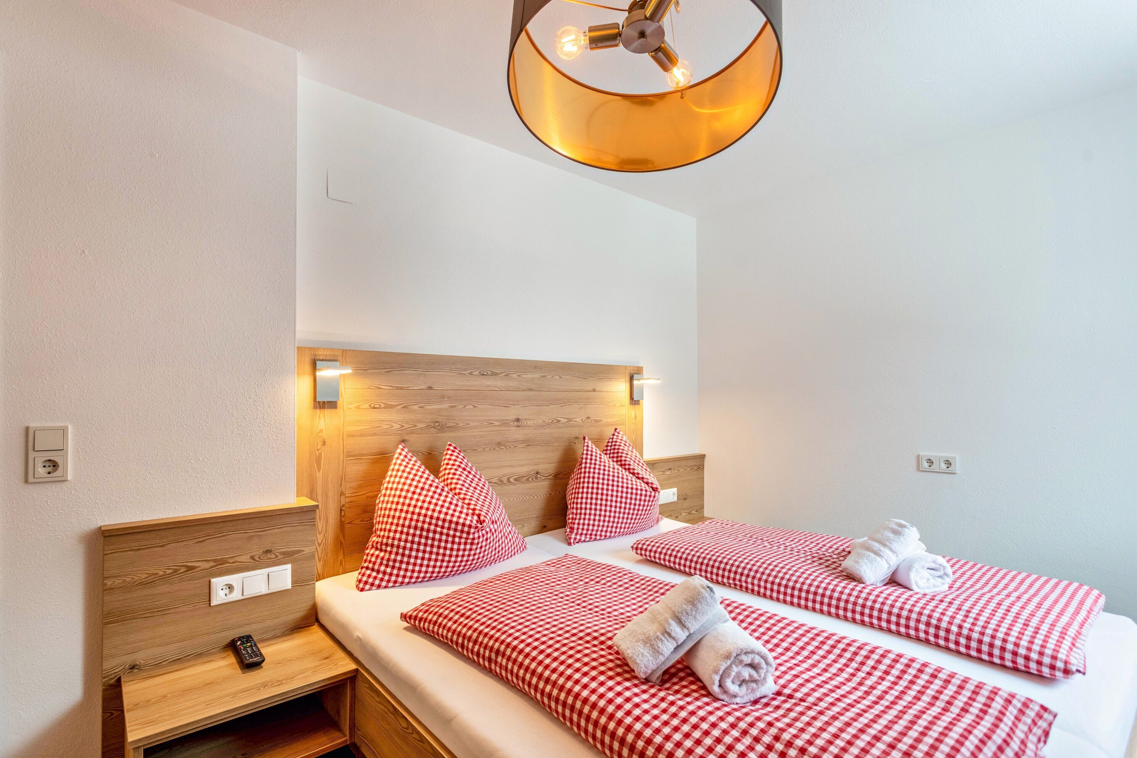 Ein Hotelzimmer mit einem Holzkopfteil, zwei Betten mit rot-weiß karierter Bettwäsche, einem kleinen Nachttisch mit Fernbedienung und einer Wandleuchte.