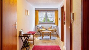 Een gang met een rood tapijt, houten deuren, een tafel met stoelen, een bijzettafeltje met tijdschriften, en een raam met gordijnen.