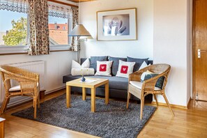 Ein Wohnzimmer mit einer Couch, zwei Korbstühlen, einem Couchtisch, einer Lampe, einem gerahmten Bild und einem Fenster mit Vorhängen.