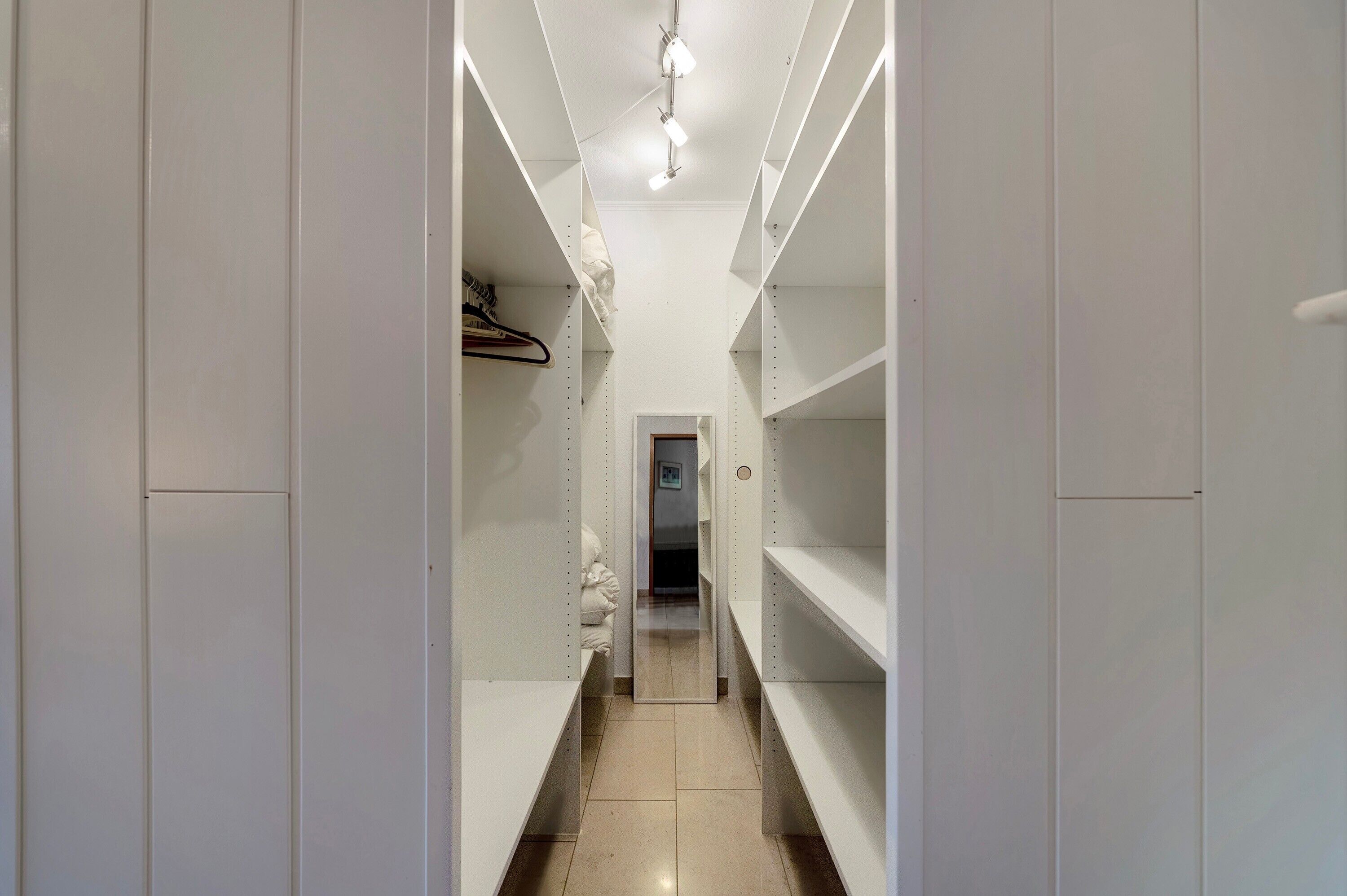 Un vestidor estrecho con estantes blancos y una barra para colgar ropa, que conduce a una puerta al final.