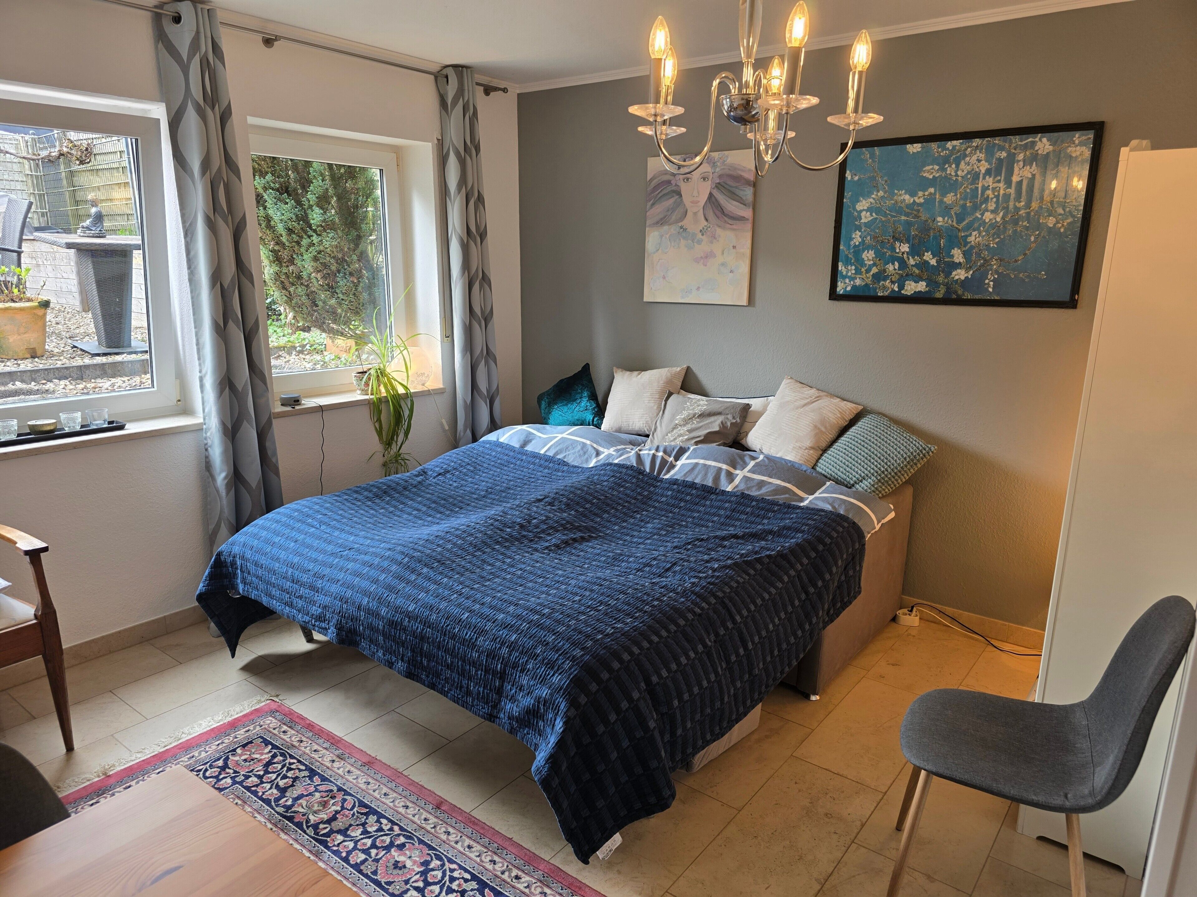 A bedroom with a bed, a chair, a window with curtains, a chandelier, and artwork on the wall.