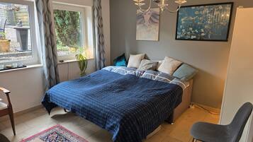 A bedroom with a bed, a chair, a window with curtains, a chandelier, and artwork on the wall.