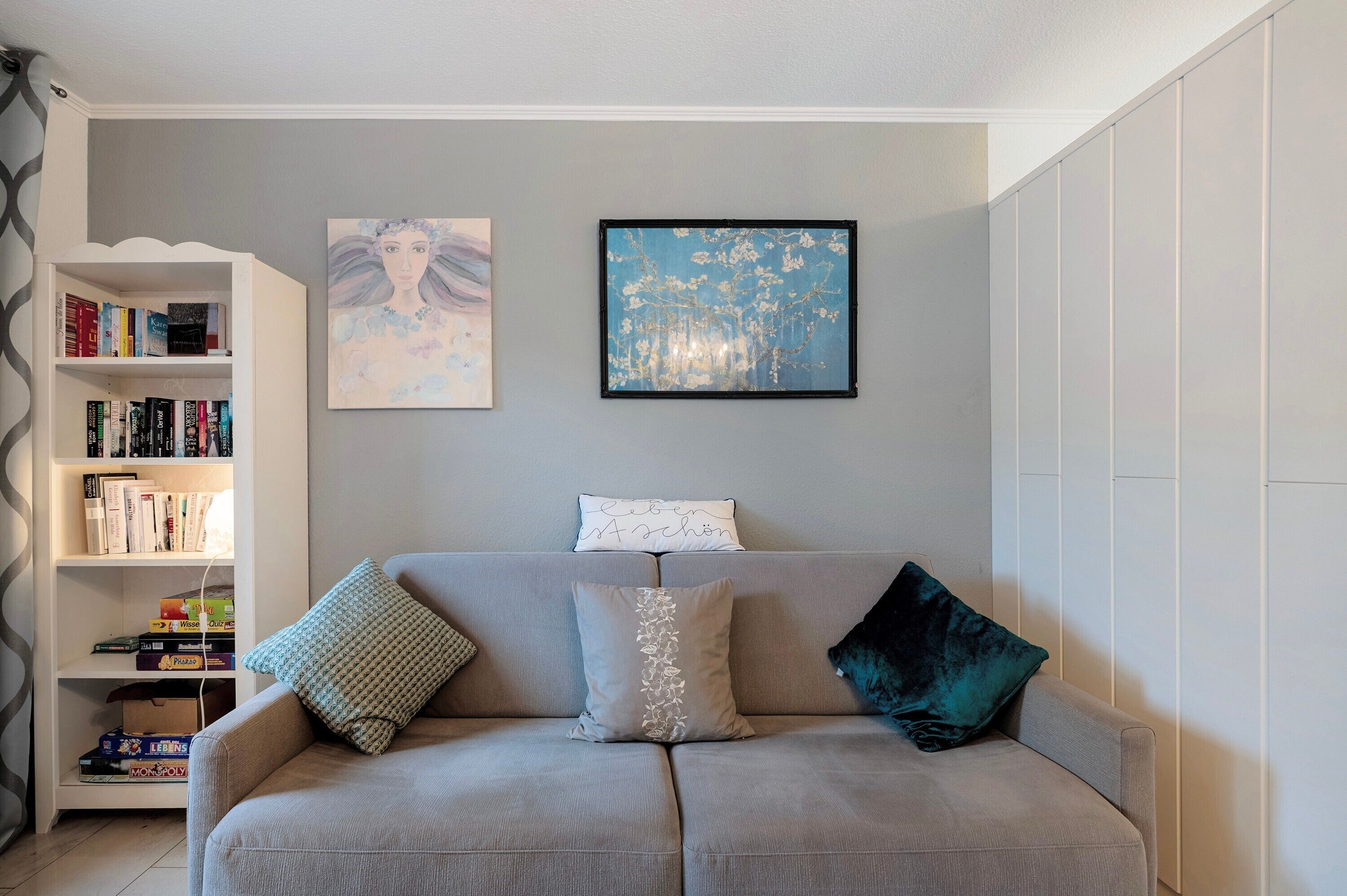 A living room with a grey sofa, a bookshelf, and two framed artworks on the wall.
