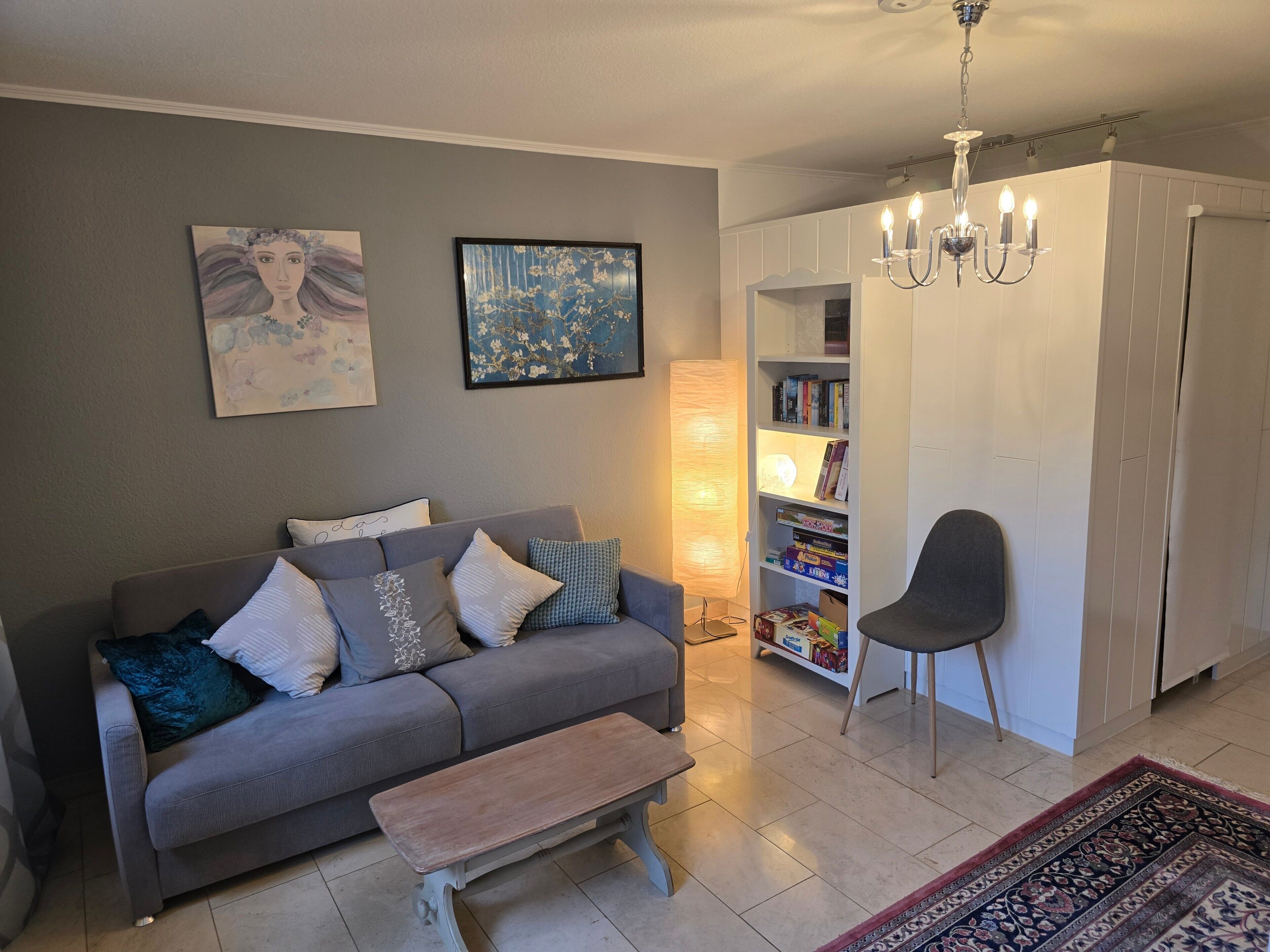 Un salon avec un canapé gris, un banc en bois, une bibliothèque, un fauteuil et un lustre.