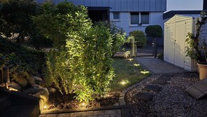 Un jardín con un sendero, un cobertizo blanco y una zona de estar con una silla tejida.