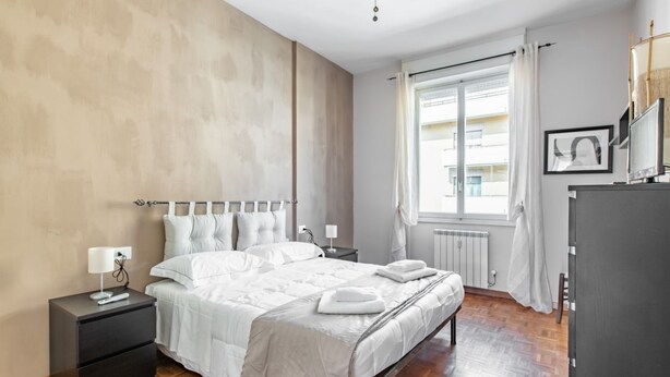 Un dormitorio con cama, mesita de noche, cómoda, ventana con cortinas y radiador.
