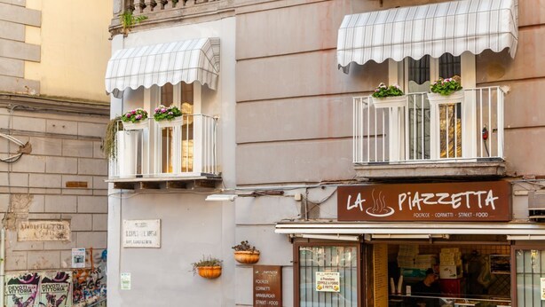 Ein Gebäude mit einer Schaufensterfront, auf der 'La Pizzetta' steht und das Essen sowie Imbisse anbietet.