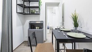 A compact kitchen with a built-in coffee machine, a dining table set for two, and a small cabinet with shelves.