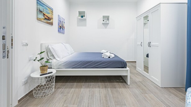 A bedroom with a bed, a wardrobe, a small bedside table with a plant, and wall-mounted shelves.