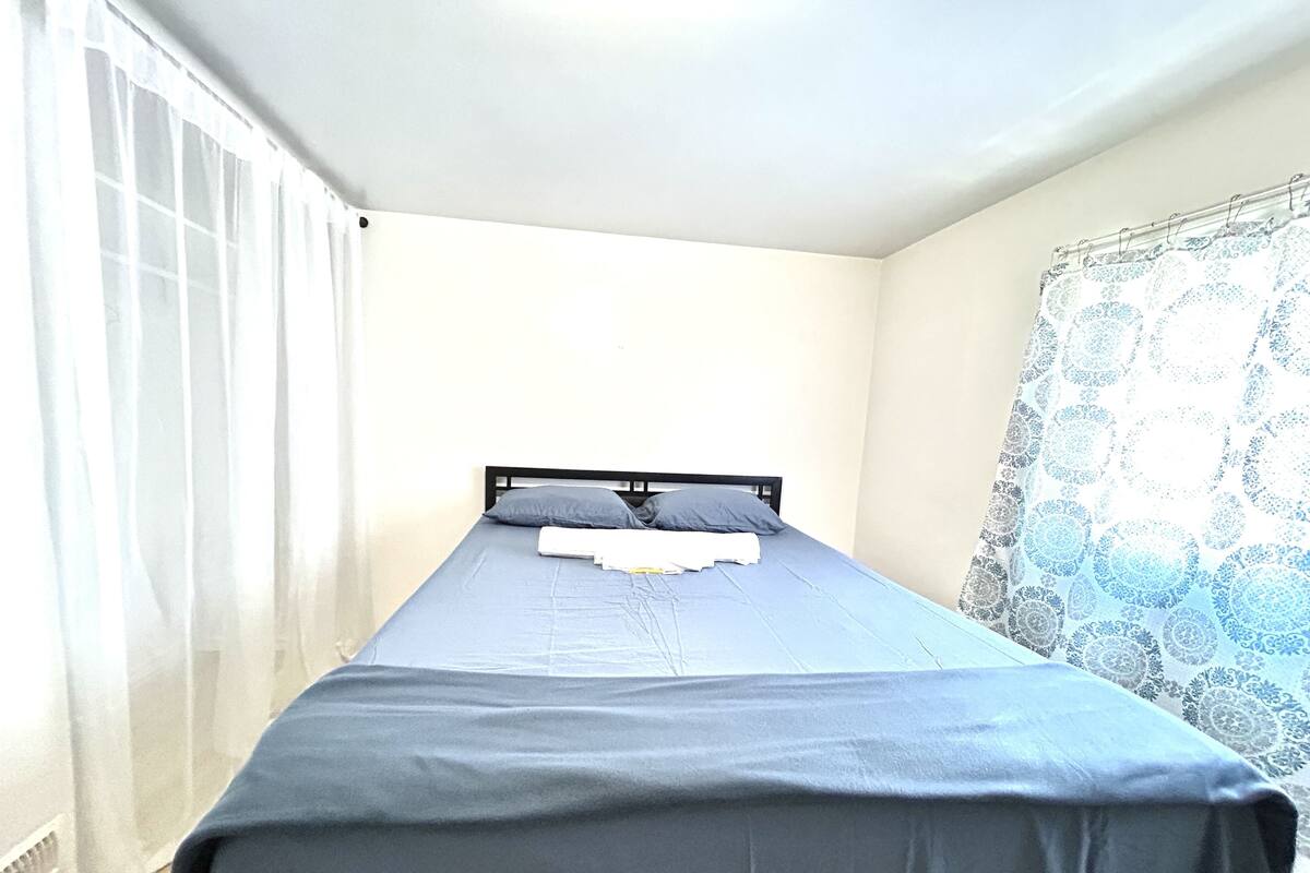 Un lit simple avec un matelas et des oreillers bleus, une couverture blanche et une fenêtre ornée de rideaux légers.