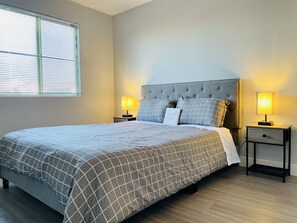 A neatly made bed with a grey checkered duvet, two bedside tables with lamps, a tufted headboard, and a window with blinds.