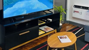 Huoneessa on suuri, litteänäyttöinen televisio, joka on sijoitettu mustalle jalustalle. Lisäksi siellä on pieni puinen pöytä, jonka päällä on lehti, ruukkukasvi ja patteri.