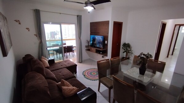 A living room with a brown sofa, a TV on a wooden shelf, a dining table with chairs, and a balcony with a view of buildings.