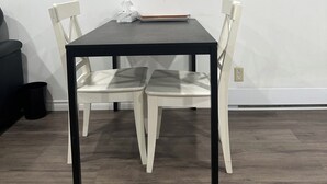 Een eetruimte met een zwarte tafel en vier witte stoelen, een bijzettafeltje met zakdoosjes, en een bloemschilderij aan de muur.