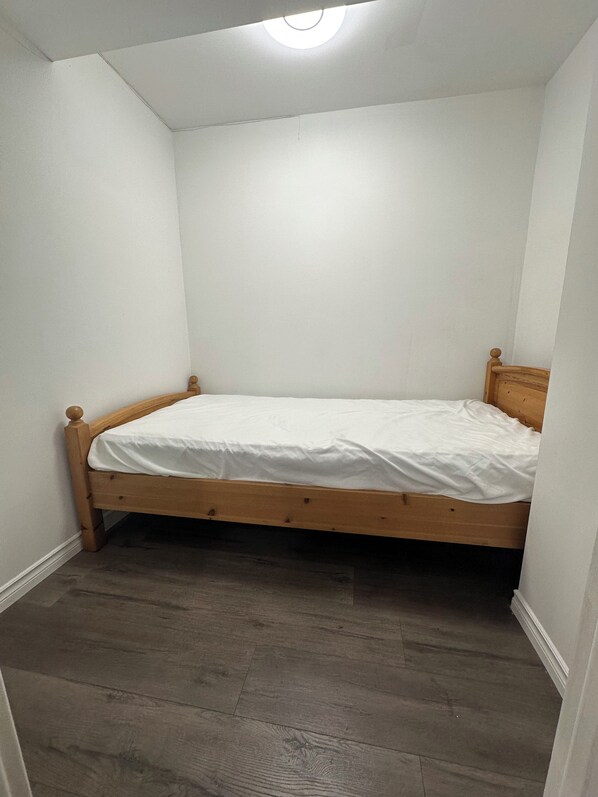 A small, single-bed room with a wooden frame, white walls, and a wooden floor.