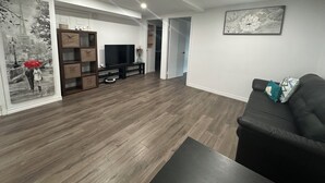 Een moderne woonkamer met een flatscreen televisie op een houten standaard, een zwarte leren bank en een salontafel. Er hangt een schilderij aan de muur en staat een houten kast met lades.