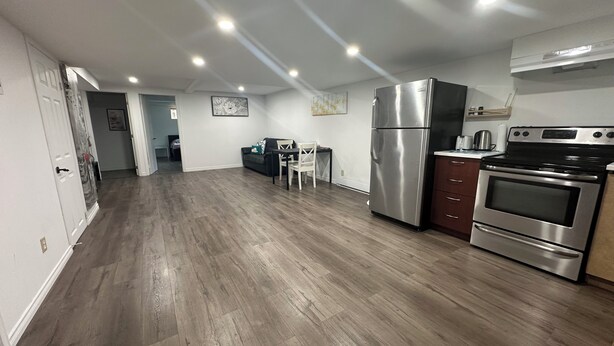 Een moderne keuken met een roestvrijstalen koelkast, oven en kookplaat. Er staat een eettafel met stoelen en een bankstel op de achtergrond.
