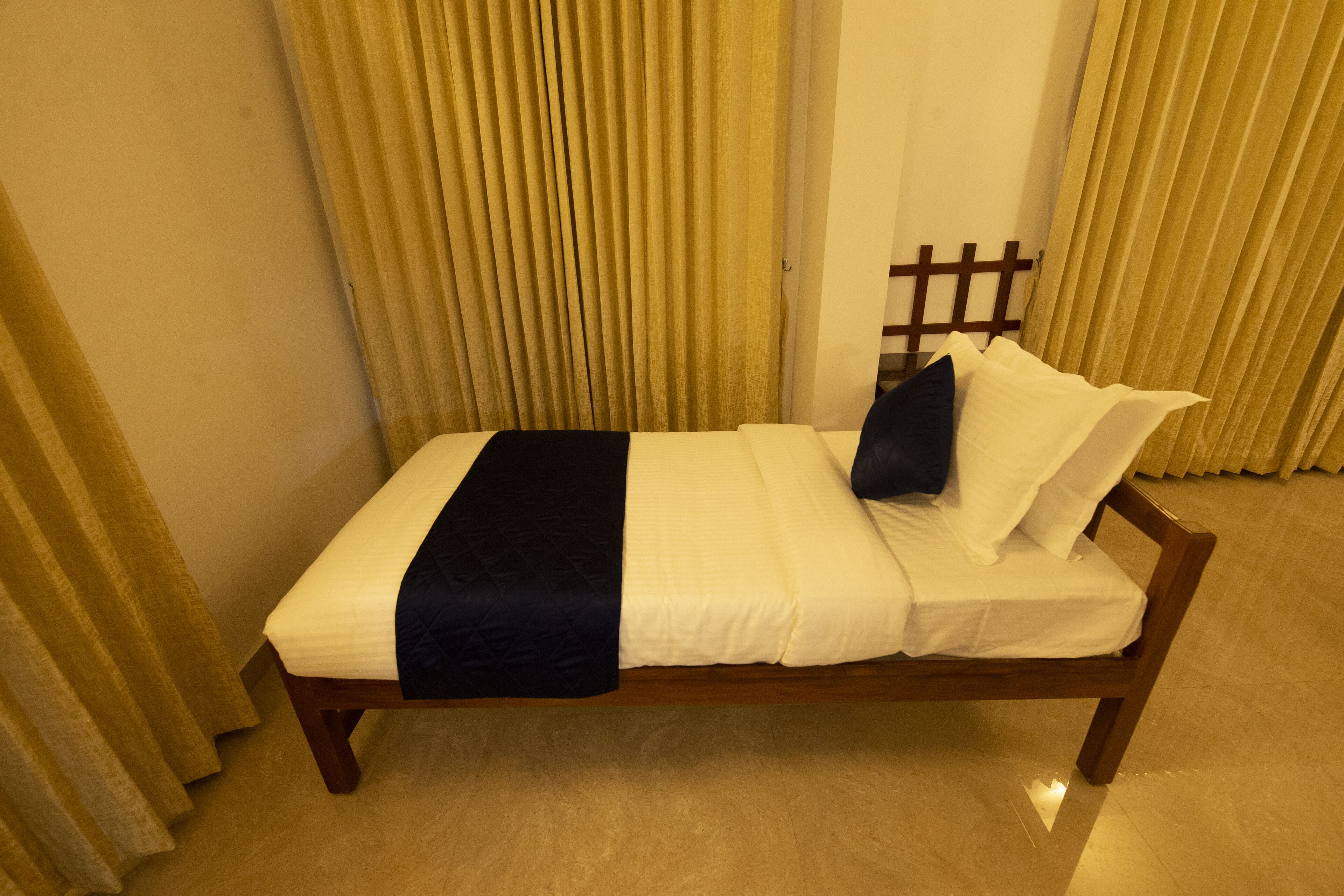 A single bed with a white mattress, a dark blue bedspread, and two pillows. The bed is positioned against a wall with a wooden headboard.