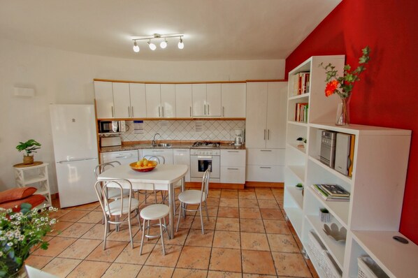 A compact kitchen with white cabinets, a dining table with chairs, a refrigerator, and a bookshelf with books and a vase of flowers.