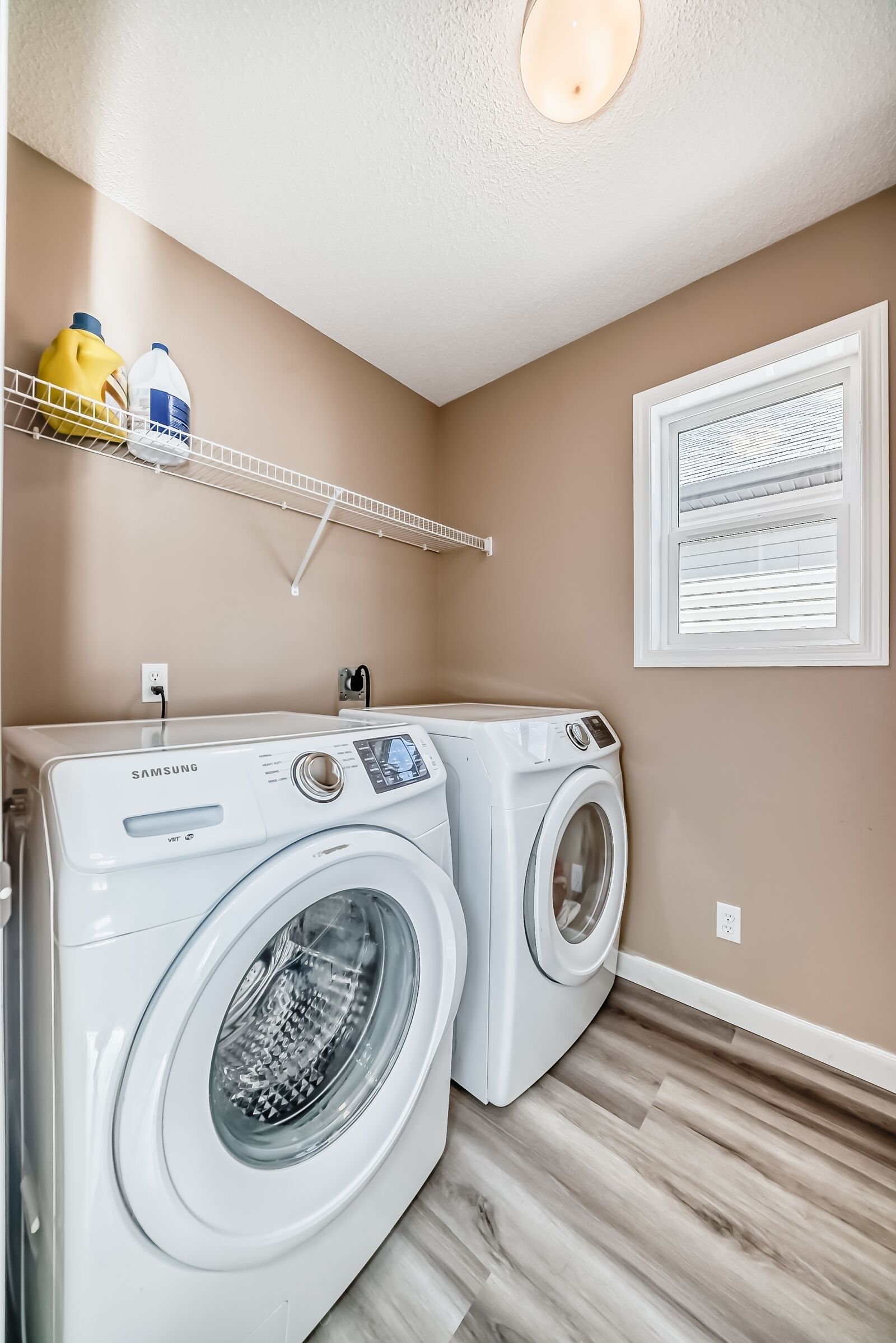 A laundry area with two Samsung front-loading washers, a shelf with detergent bottles, a window with blinds, and a light fixture on the ceiling.