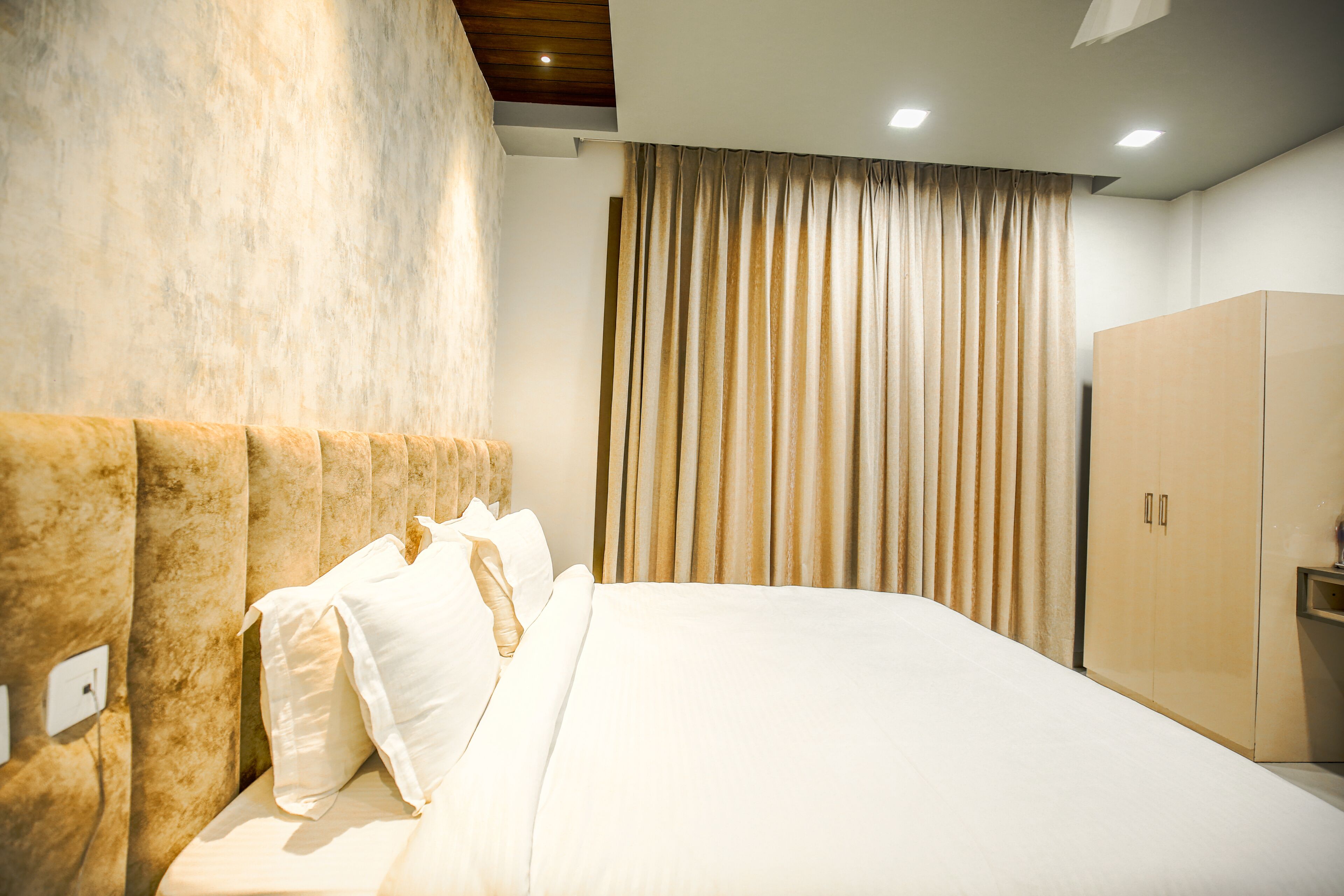 A neatly made bed with white linens and a wooden headboard, a wardrobe, and a large window with curtains.