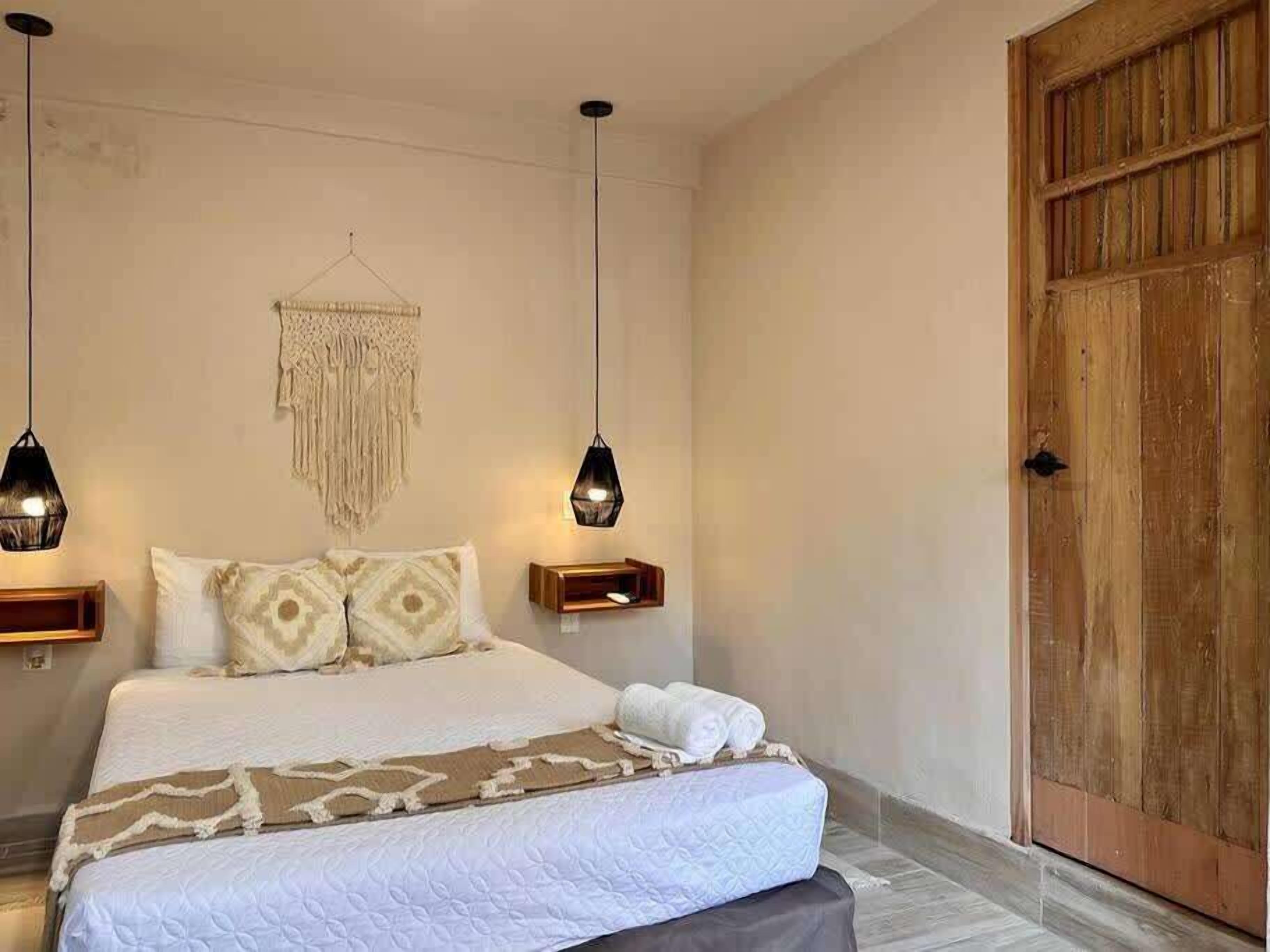 Une chambre à coucher avec un lit, deux tables de chevet, une décoration murale en macramé et une porte en bois.