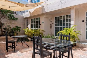 An outdoor patio with a glass table and chairs, surrounded by potted plants and a beige awning.