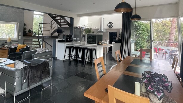 Eine moderne Küche mit einer zentralen Kochinsel, ein Essbereich mit Holztisch und -stühlen sowie eine Treppe, die in ein Obergeschoss führt.